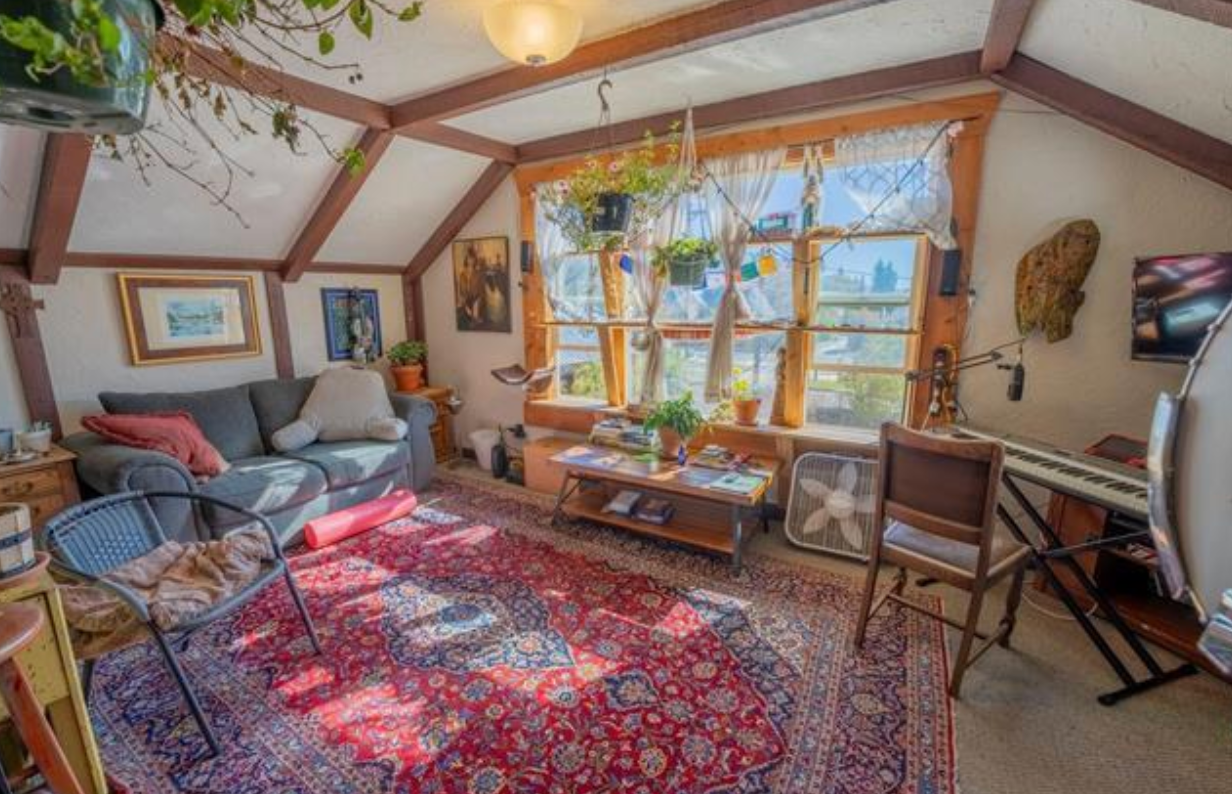 Cozy sunroom with large windows, red patterned rug, sofa, chairs, and a coffee table with plants and artwork.