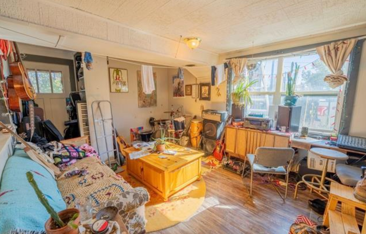 Cluttered sunlit living room with sofa, wooden coffee table, desk, chair, and large windows