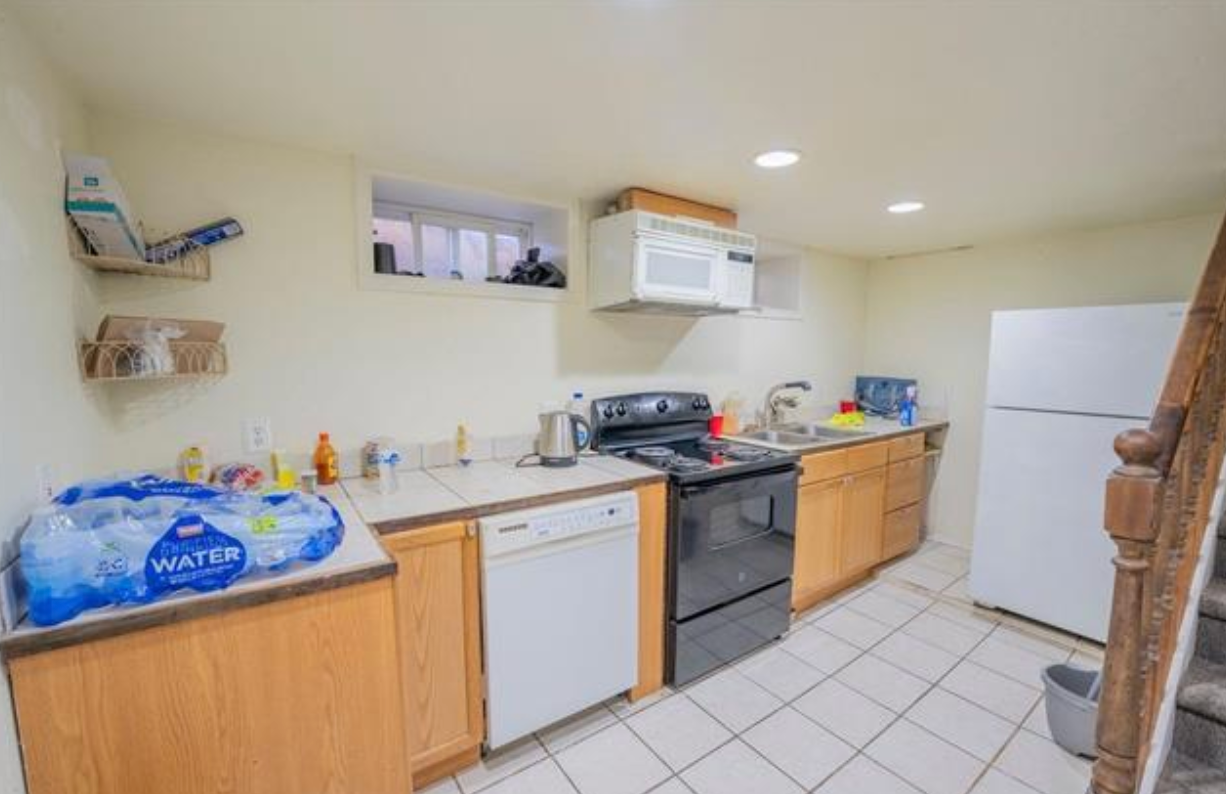 Small kitchen with wood cabinets, white appliances, tiled floor, and a water jug on the counter.