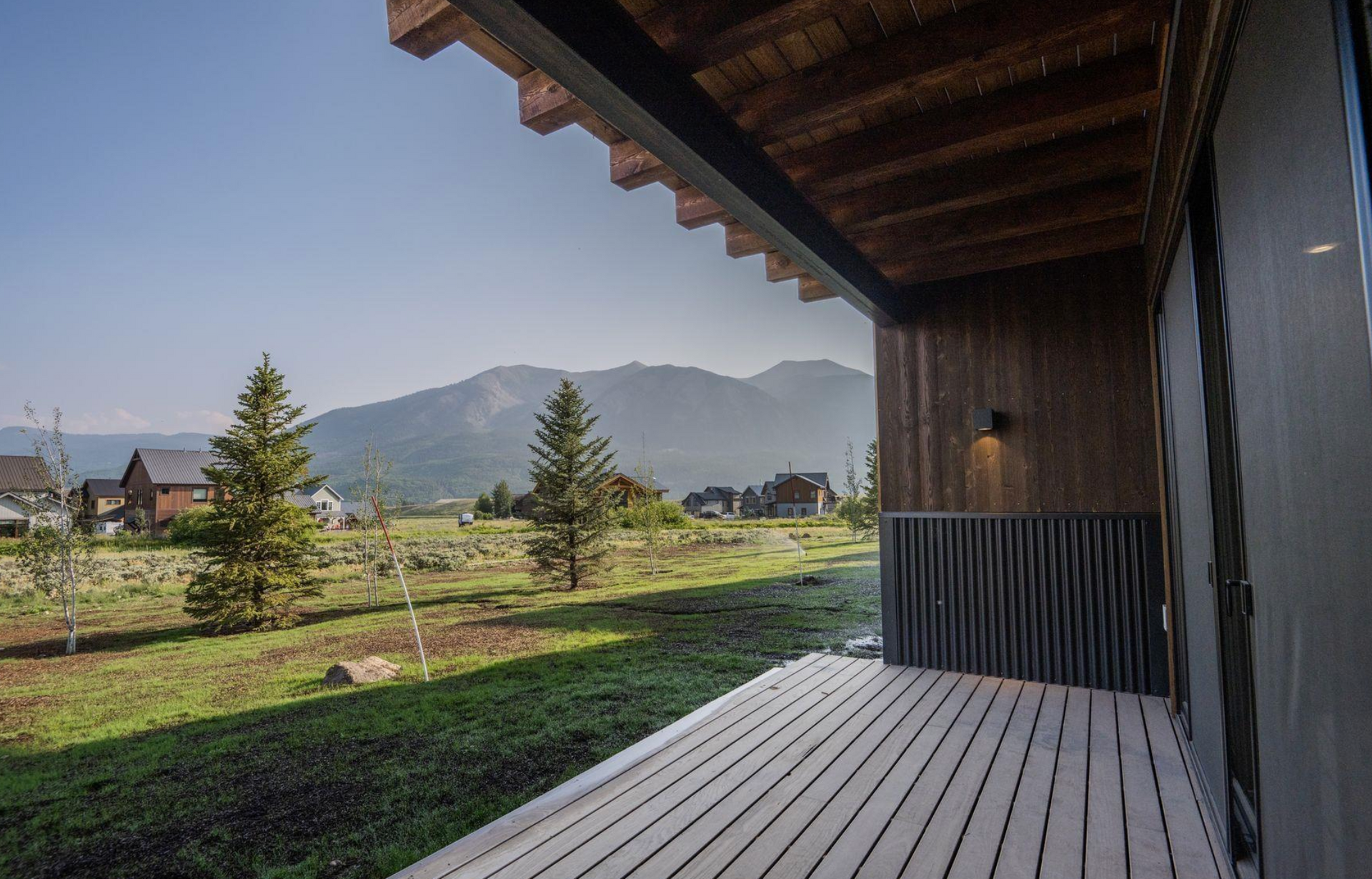 Wooden patio overlooking a grassy yard, pine trees, and distant mountains under a clear sky