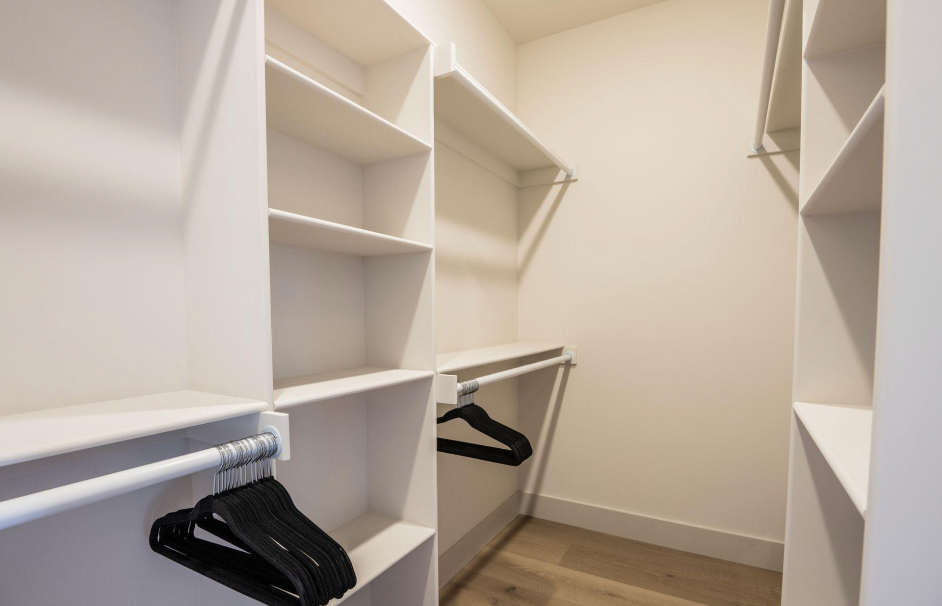 Empty walk-in closet with white shelves and hanging rods on both sides, black hangers, and wood flooring.