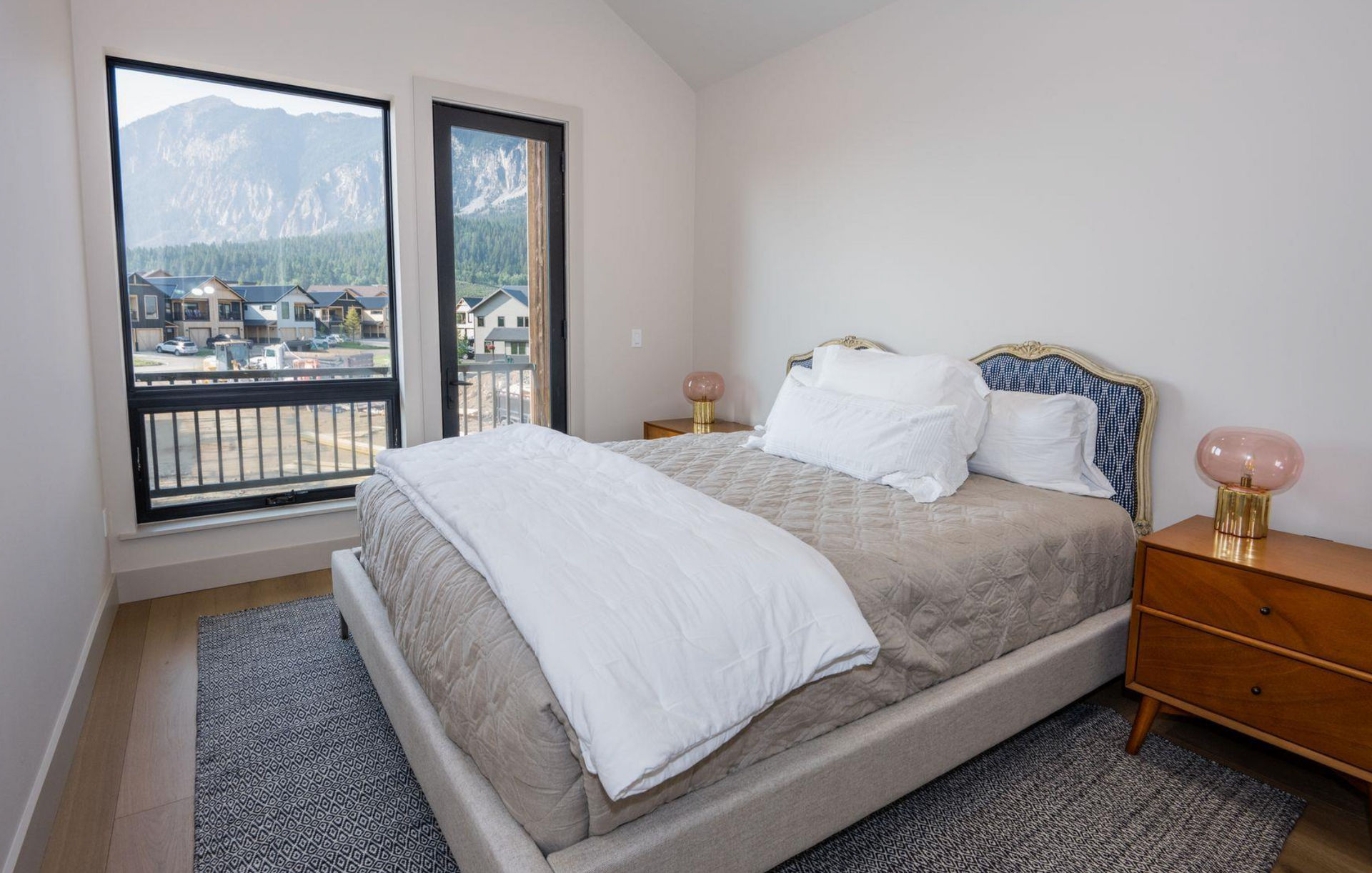 Bright bedroom with a made bed, nightstand lamps, and a balcony view of mountains and water.