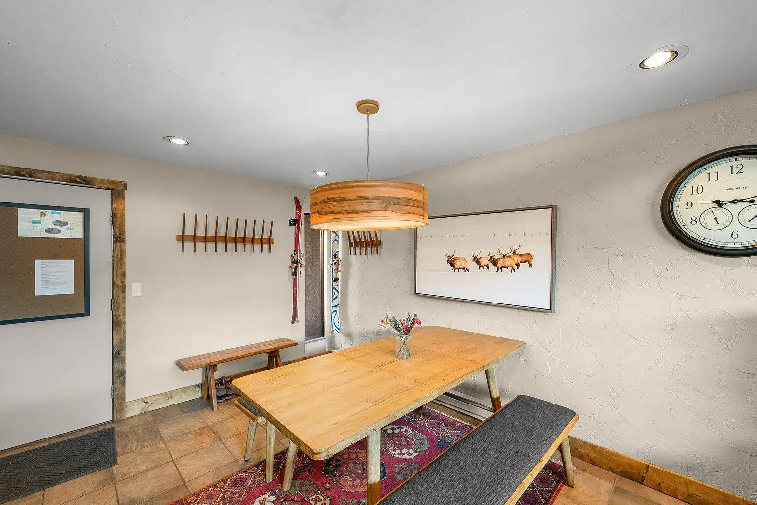 Cozy dining room with wooden table, benches, wall art, and large clock on a beige wall.