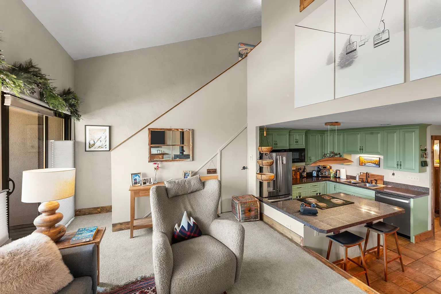 Bright living room with staircase, gray armchair, and open view into a green-and-wood kitchen
