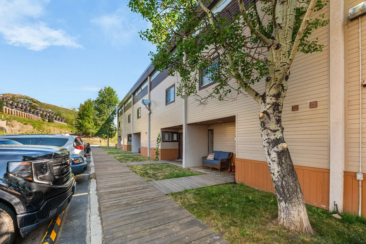 Apartment building walkway with parked cars, trees, and a grassy courtyard on a sunny day