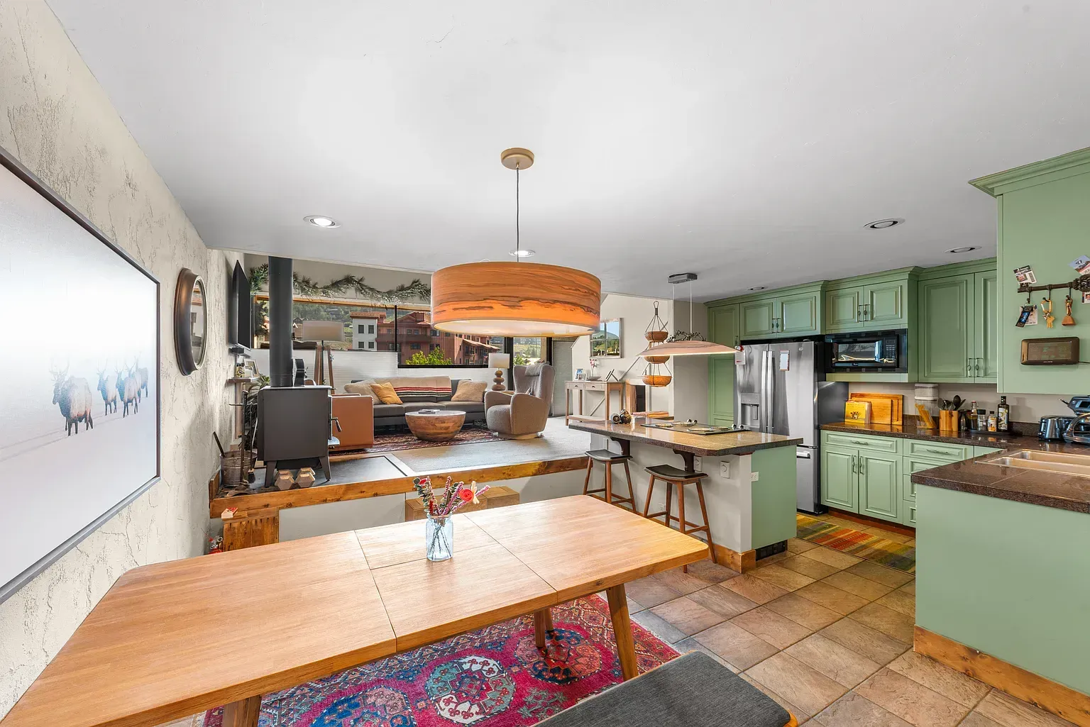 Bright open kitchen and dining area with green cabinets, wood table, and orange pendant light.