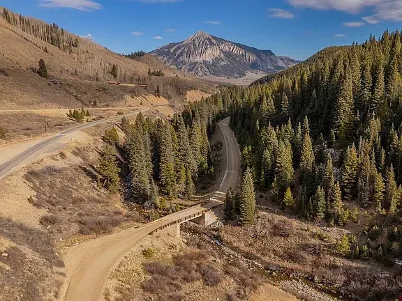 Mountain valley with a winding dirt road, pine forest, and rocky peaks under a blue sky