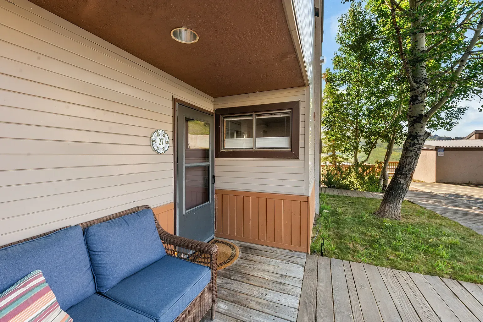 Covered porch with blue cushioned chair, screened door, and wooden deck beside a grassy yard