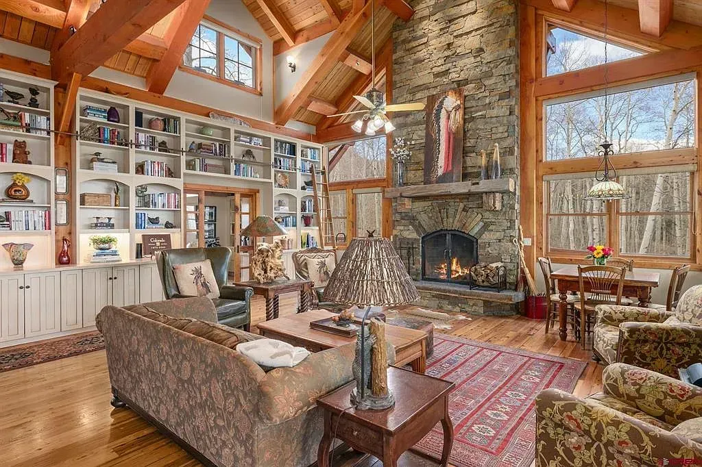Rustic living room with stone fireplace, wood beams, bookshelves, and patterned sofas in warm sunlight