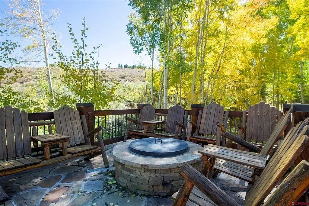 Wooden deck with Adirondack chairs вокруг a round fire pit, surrounded by yellow autumn trees and water views