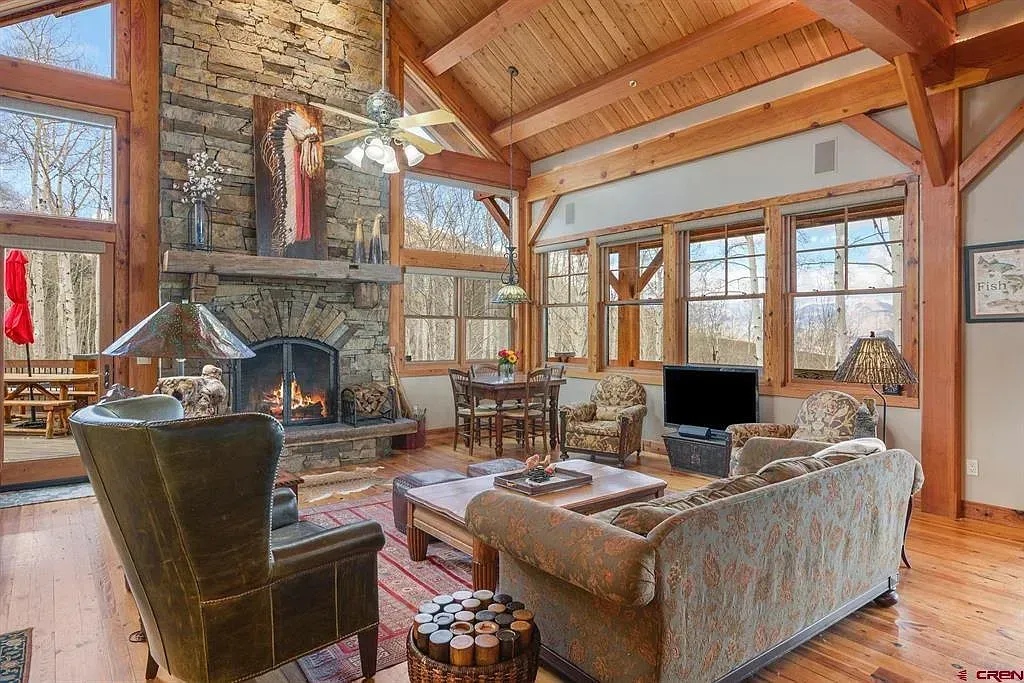 Cozy log cabin living room with stone fireplace, wood beams, large windows, and rustic furniture
