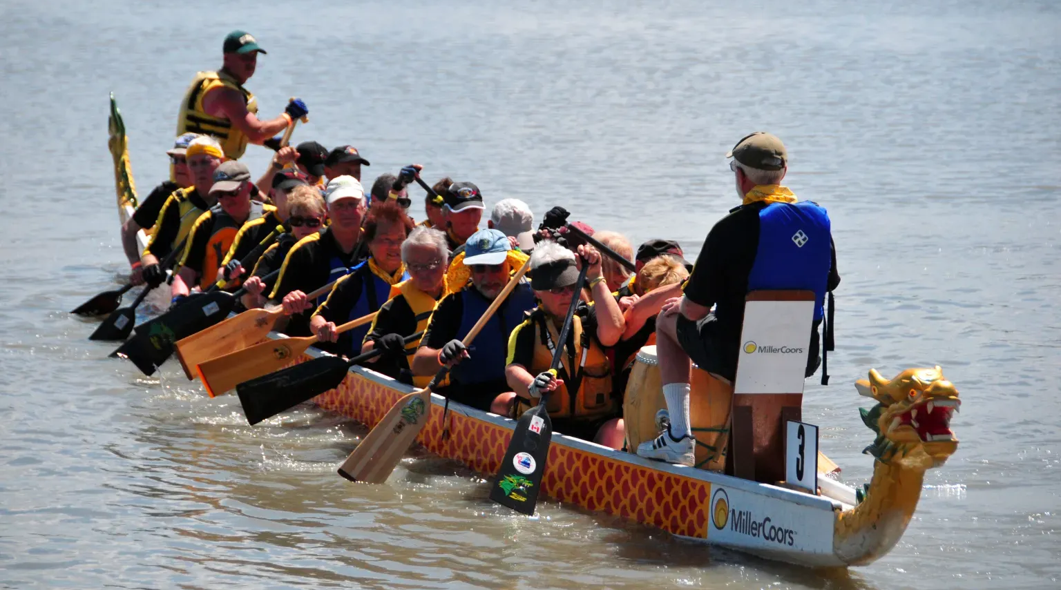 Team paddling a dragon boat on calm water, with a drummer at the front.