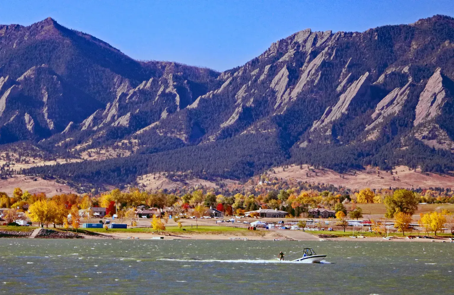 Mountain range behind a windy lakeside with autumn trees and a small boat on the water.