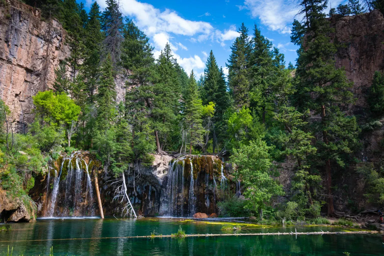 Waterfall cascading into a turquoise lake beside a rocky cliff and pine forest under a blue sky