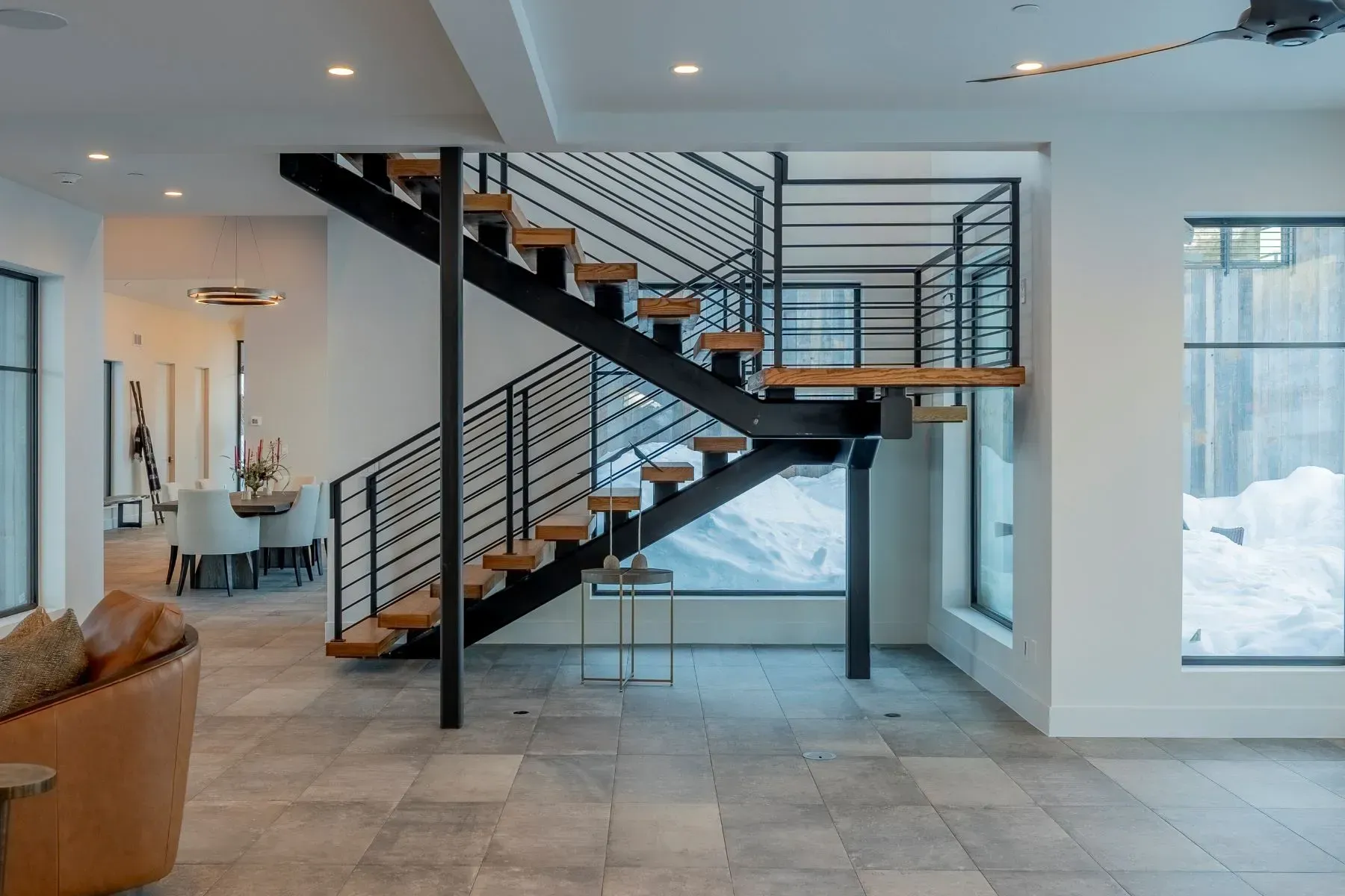 Modern open-concept interior with floating staircase, black railings, tiled floor, and large windows