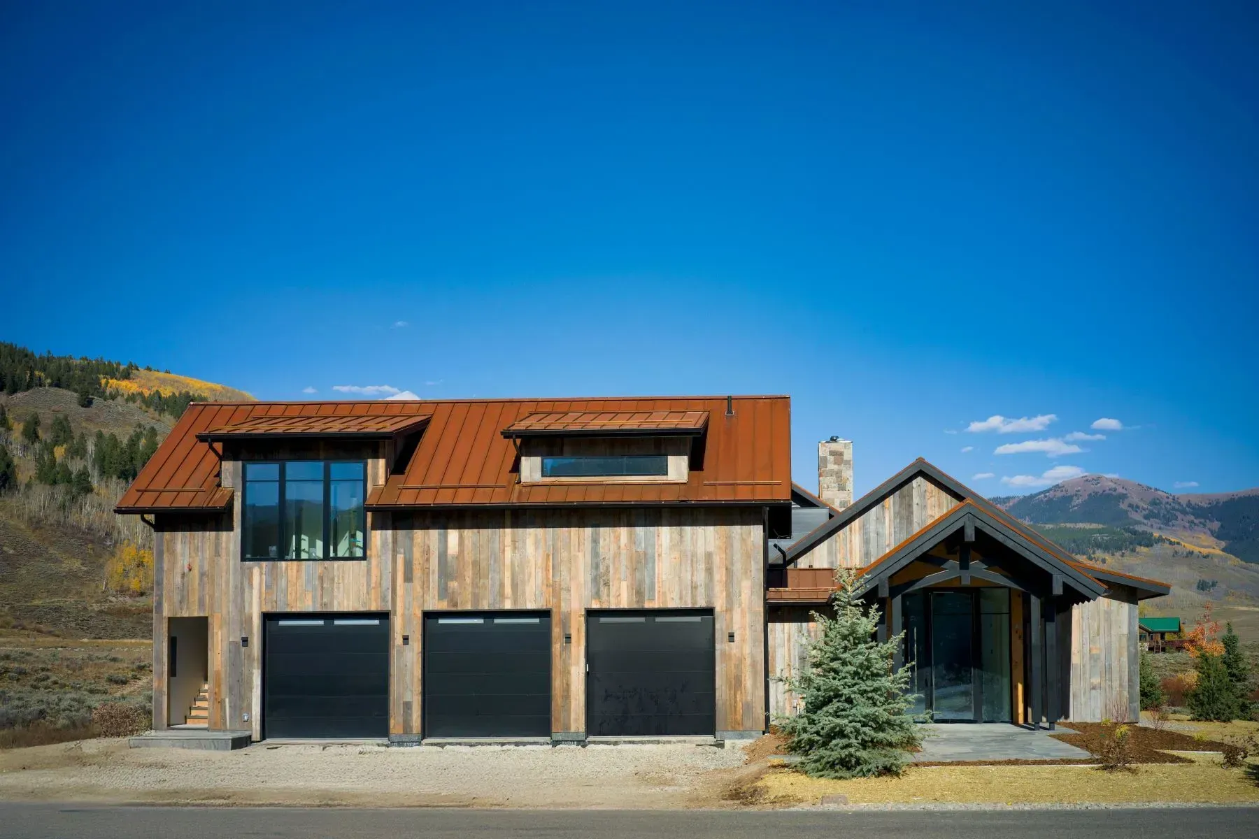 Modern house with wood and stone exterior, three garage doors, and mountain backdrop under a blue sky