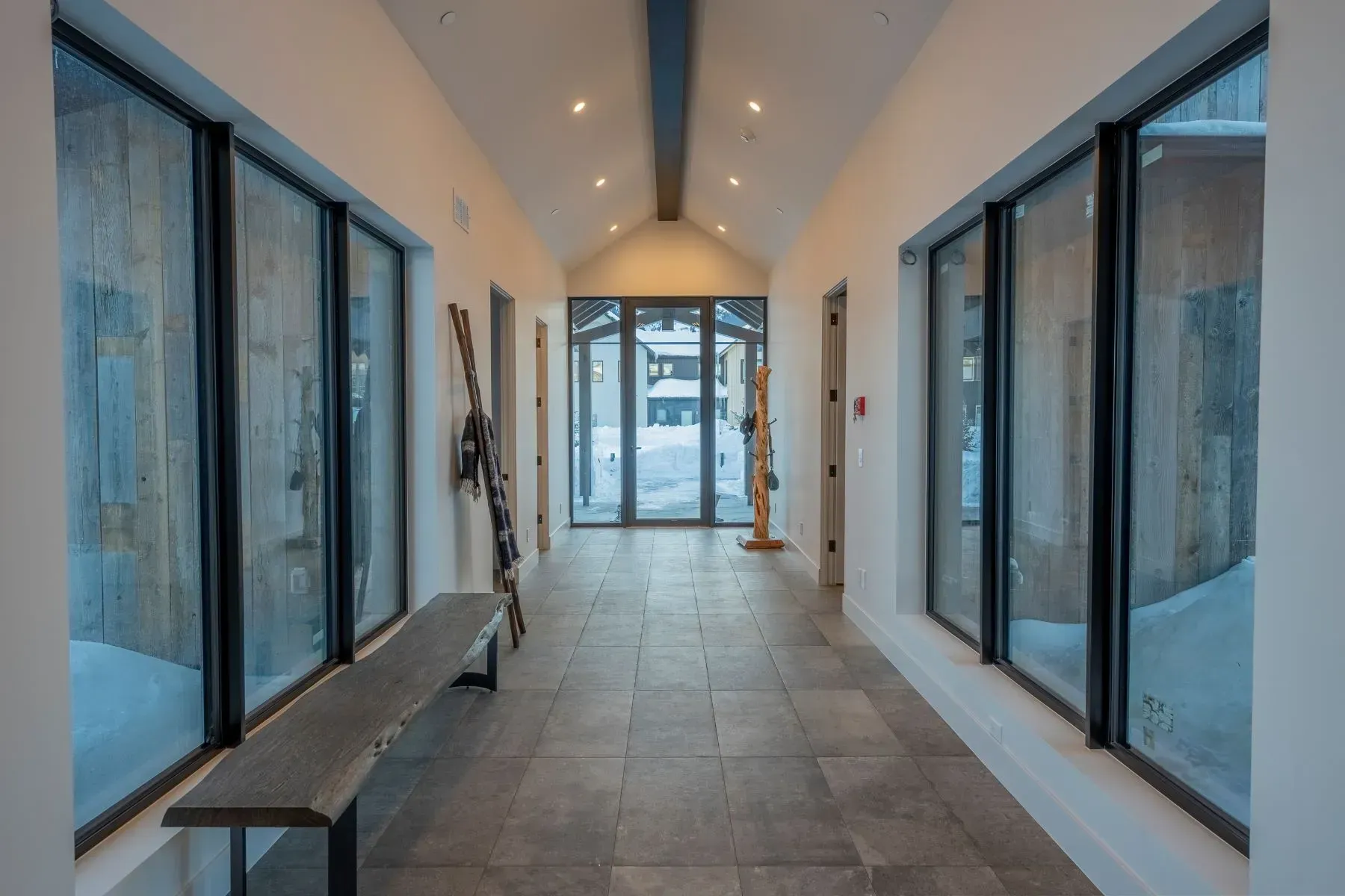 Bright modern hallway with large windows, tiled floor, bench, and glass doors at the far end