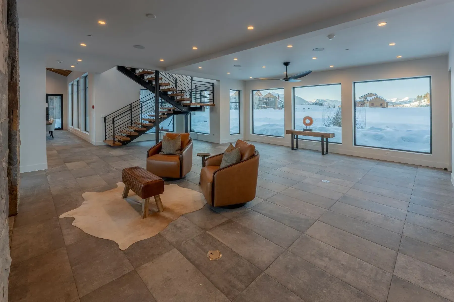 Modern open-concept living room with stone floor, leather chairs, staircase, and large windows overlooking water