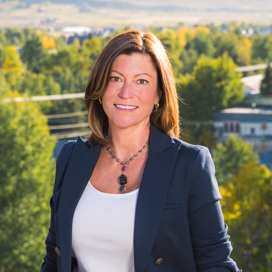 Smiling woman in navy blazer and white top standing outdoors with trees and distant hills behind her