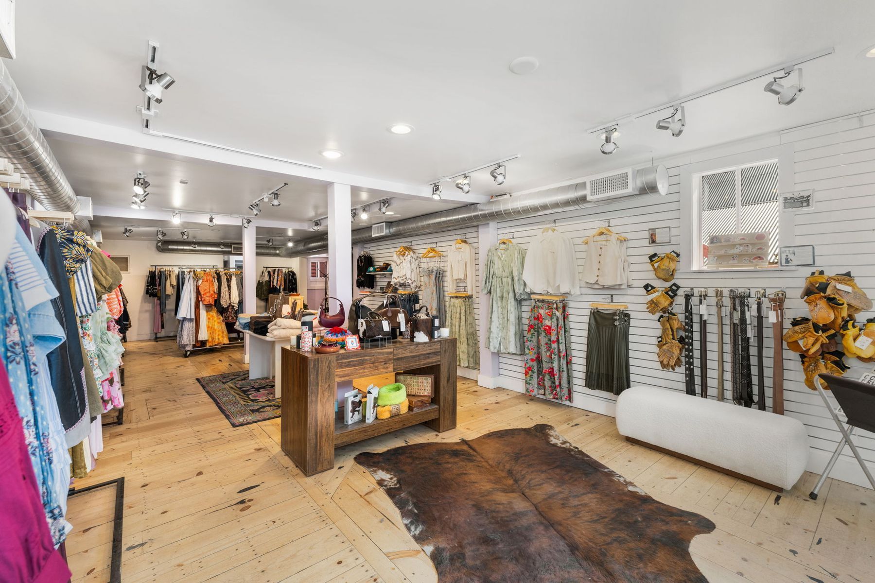 Bright boutique clothing store with racks of colorful garments, wooden display table, and patterned rug on a white floor