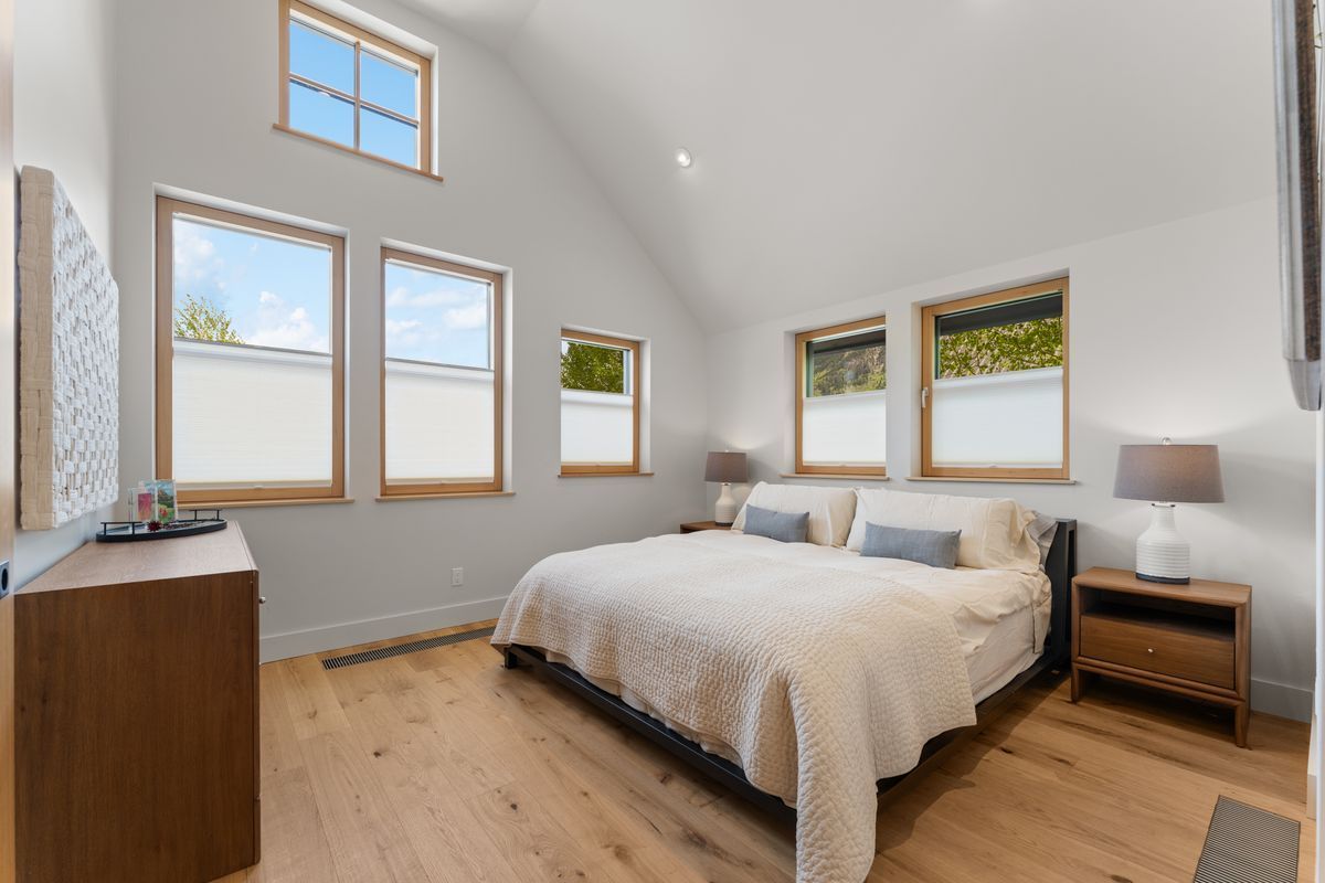 Bright bedroom with vaulted ceiling, large windows, a bed, nightstand, dresser, and wood flooring.