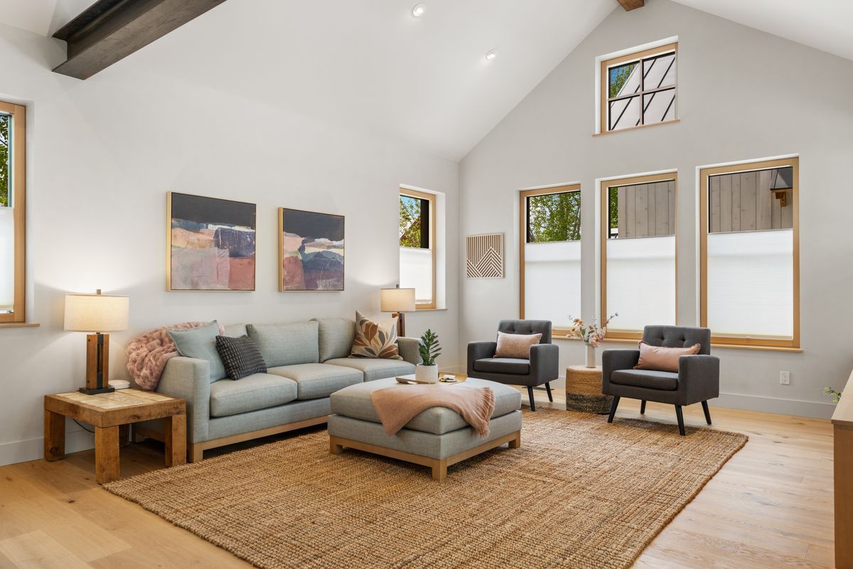 Bright living room with a gray sofa, armchairs, ottoman, woven rug, and gold-framed windows