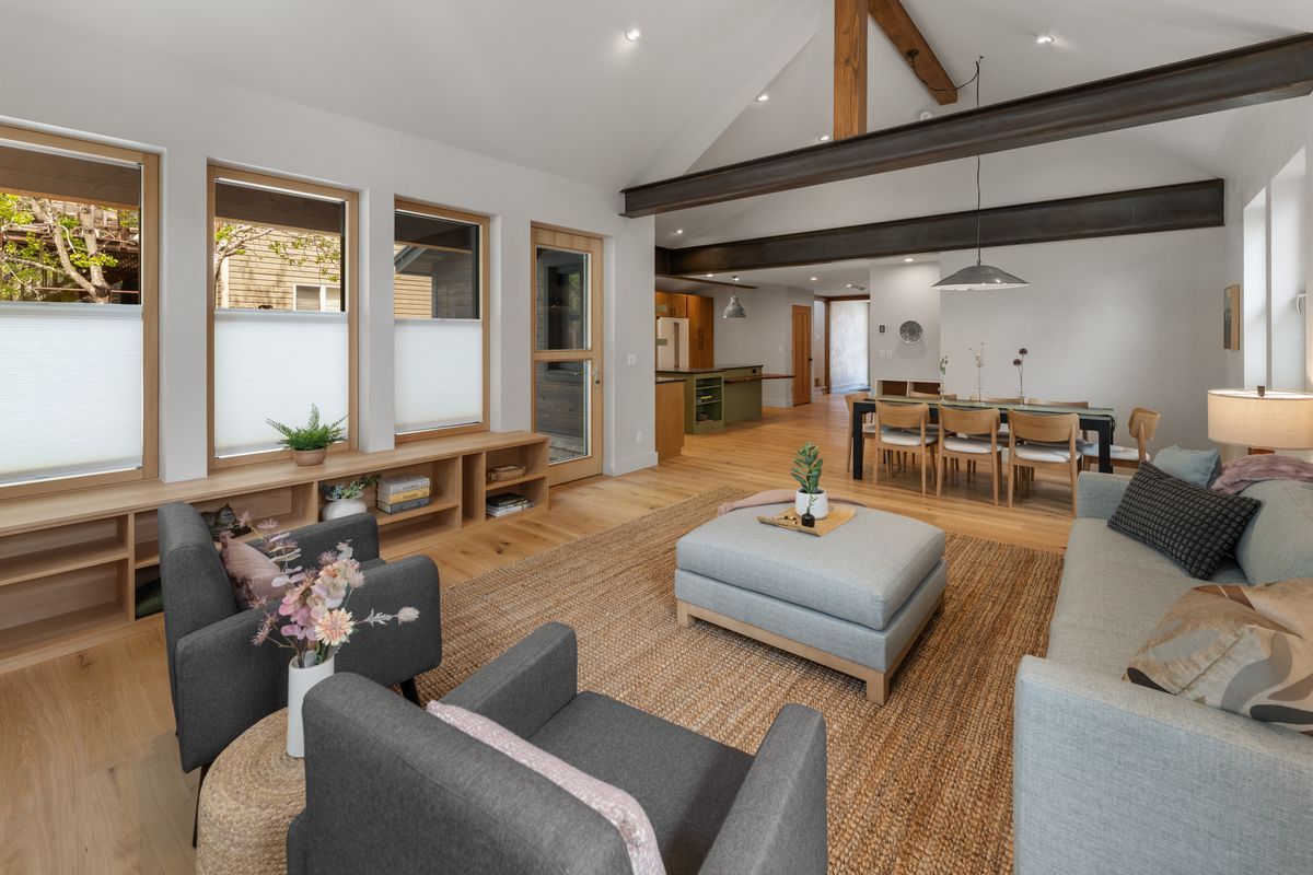 Bright open-plan living room with gray sofas, ottoman, wooden beams, and dining area in the background
