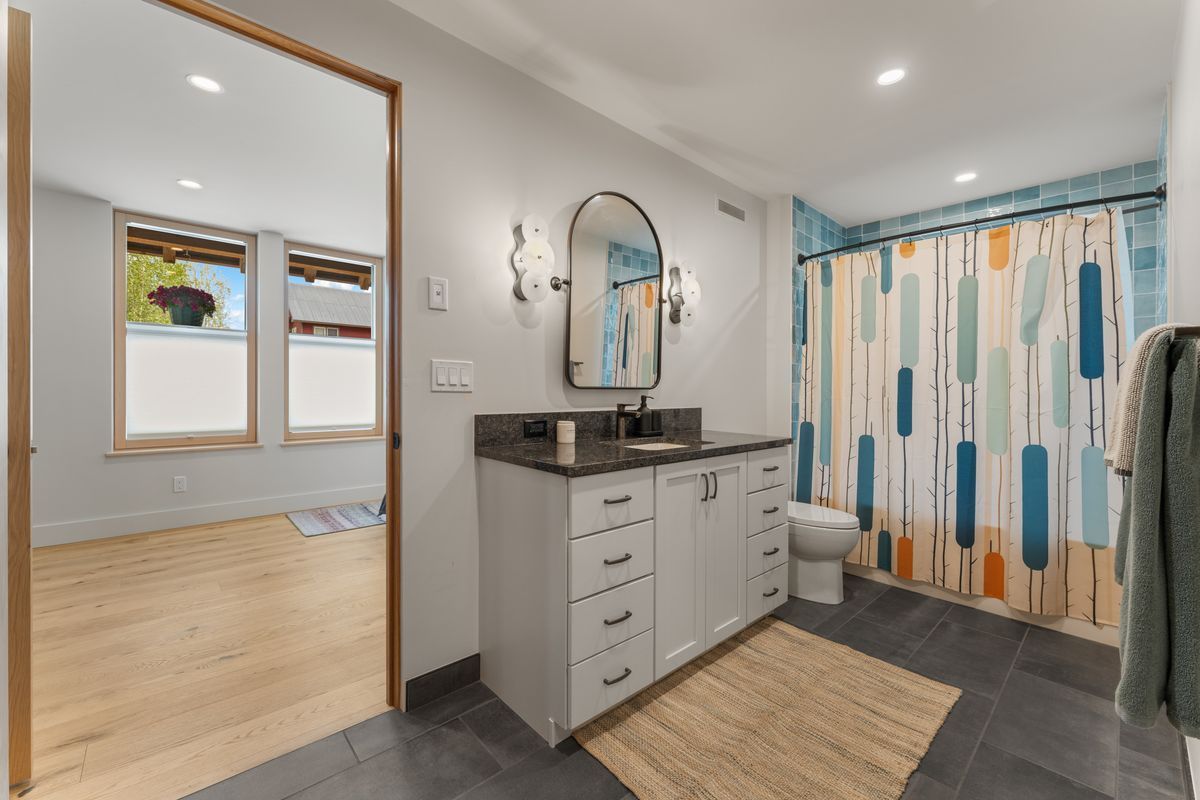 Bright bathroom with white vanity, black countertop, oval mirror, gray floor, and blue-and-orange shower curtain.