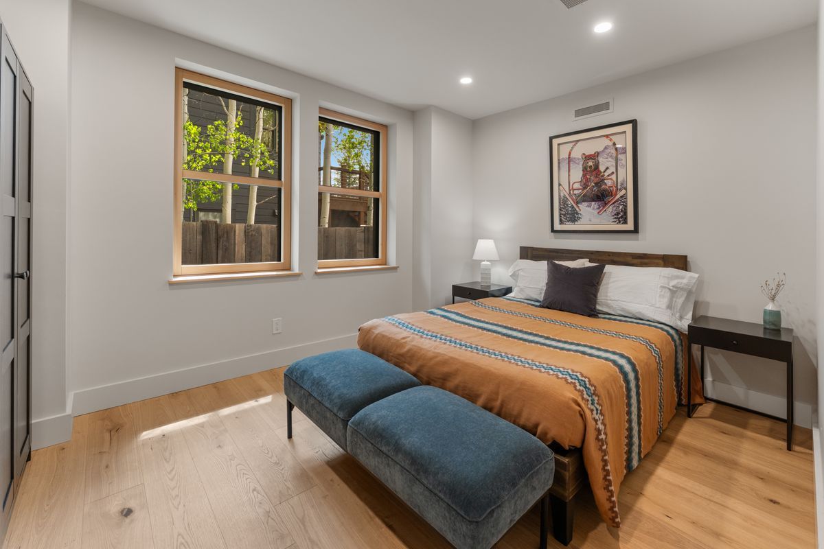 Small bedroom with a bed, blue bench, framed wall art, and two windows letting in daylight
