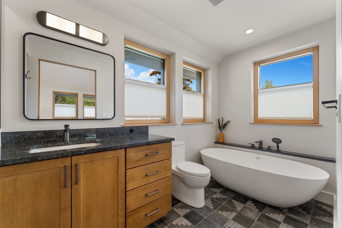 Modern bathroom with wood vanity, mirror, freestanding tub, toilet, and three windows