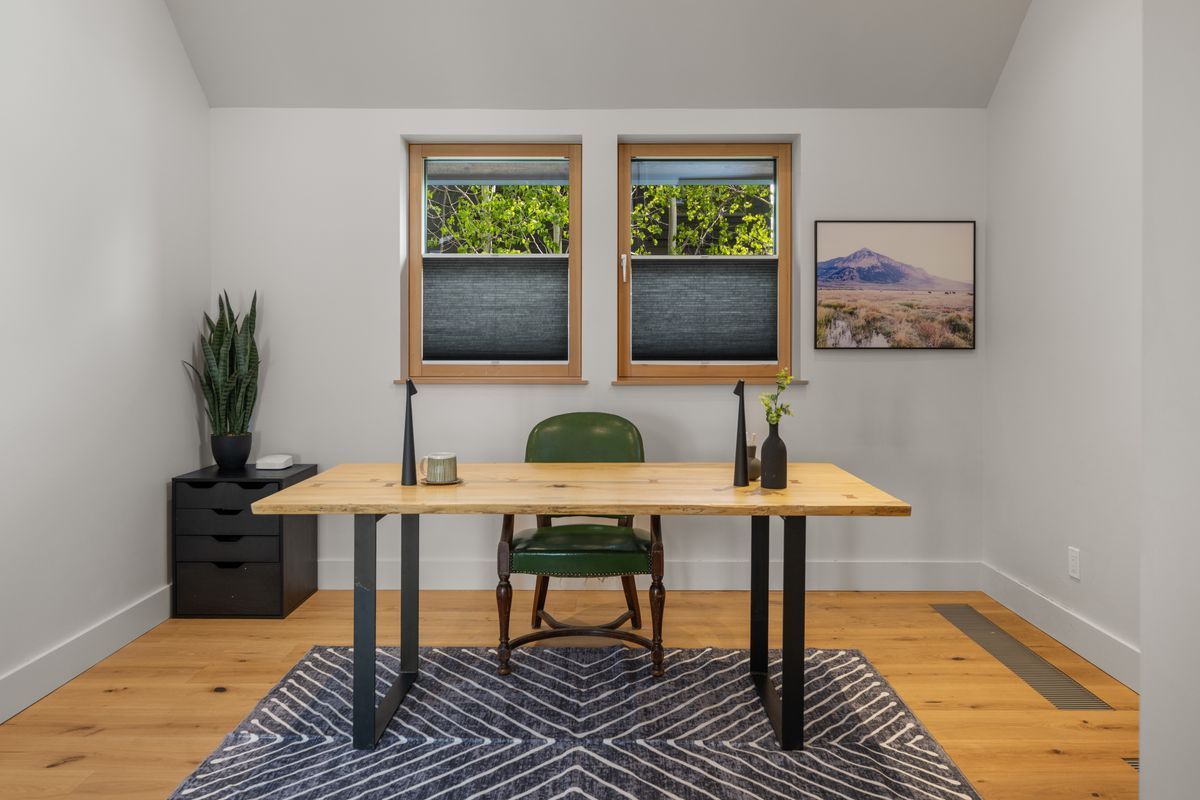 Minimal home office with wooden desk, green chair, black cabinets, plants, and two windows