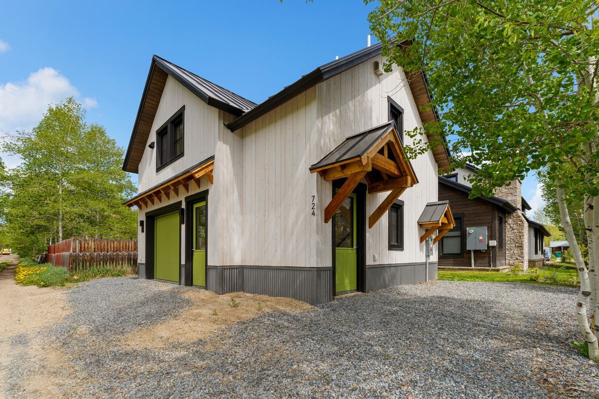 White barn-style house with dark trim and green doors on a gravel lot under a blue sky