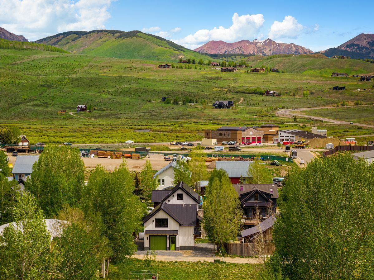 Green hillside town with houses, trees, and mountains under a blue sky