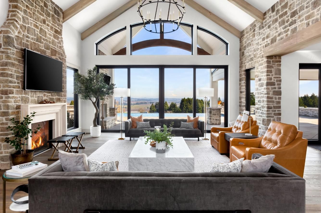 Bright modern living room with vaulted ceiling, stone fireplace, large windows, and tan leather chairs