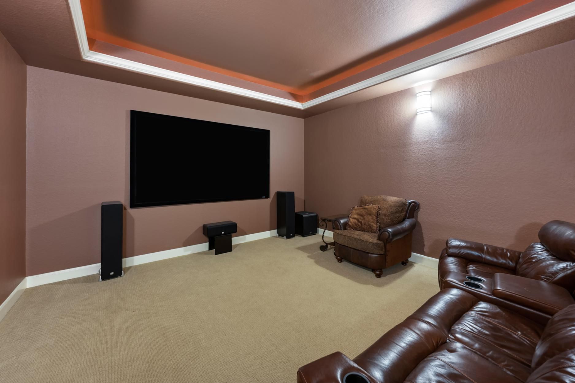 Home theater room with brown leather seating, large wall TV, and speakers on beige carpet.