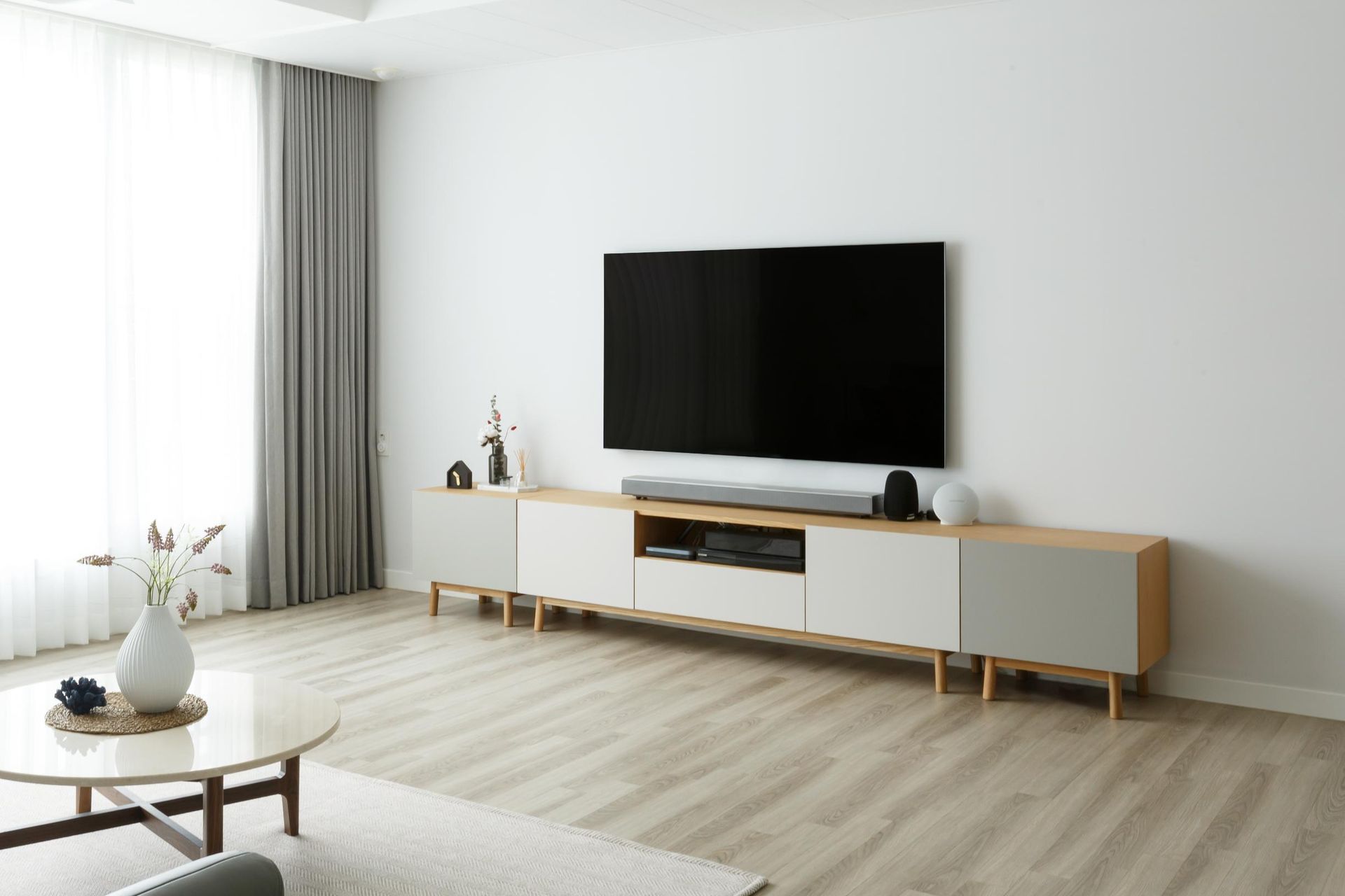 Bright minimalist living room with wall-mounted TV, white console, gray curtains, and light wood floor.