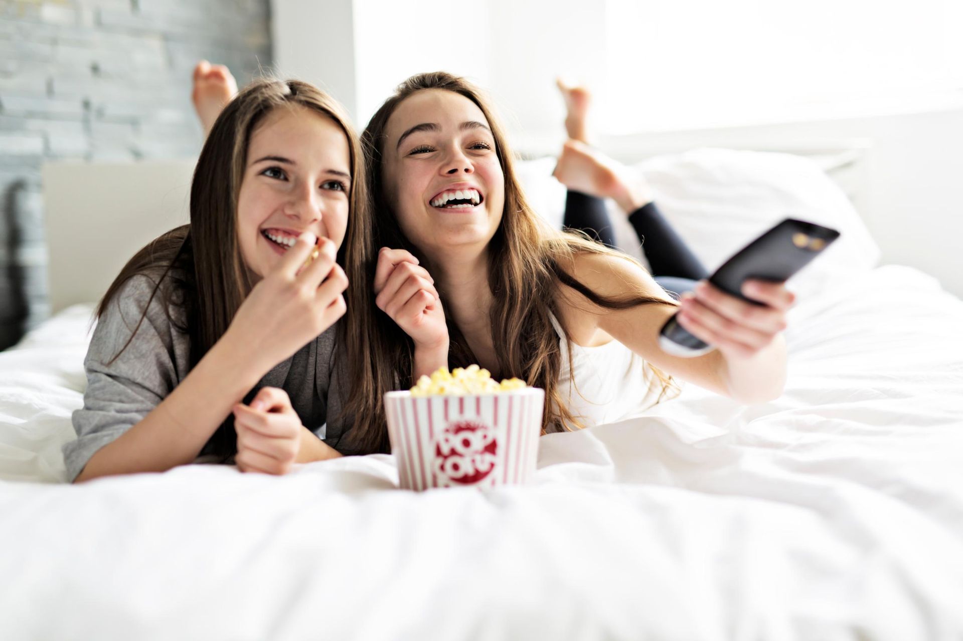Two women smiling on a bed, sharing popcorn and watching a phone with a cozy movie night scene