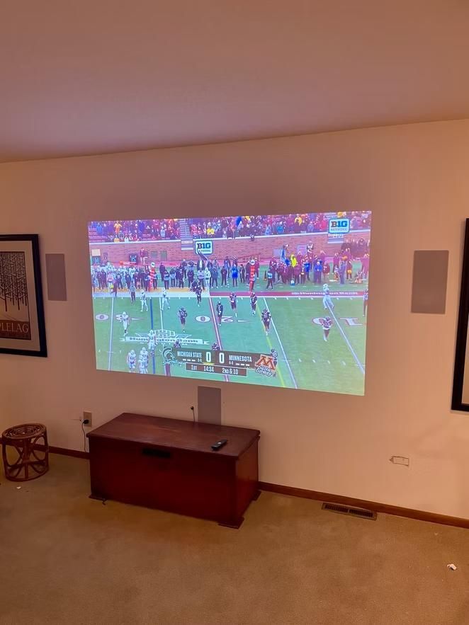 Living room with football game projected on the wall above a wooden chest and beige carpet