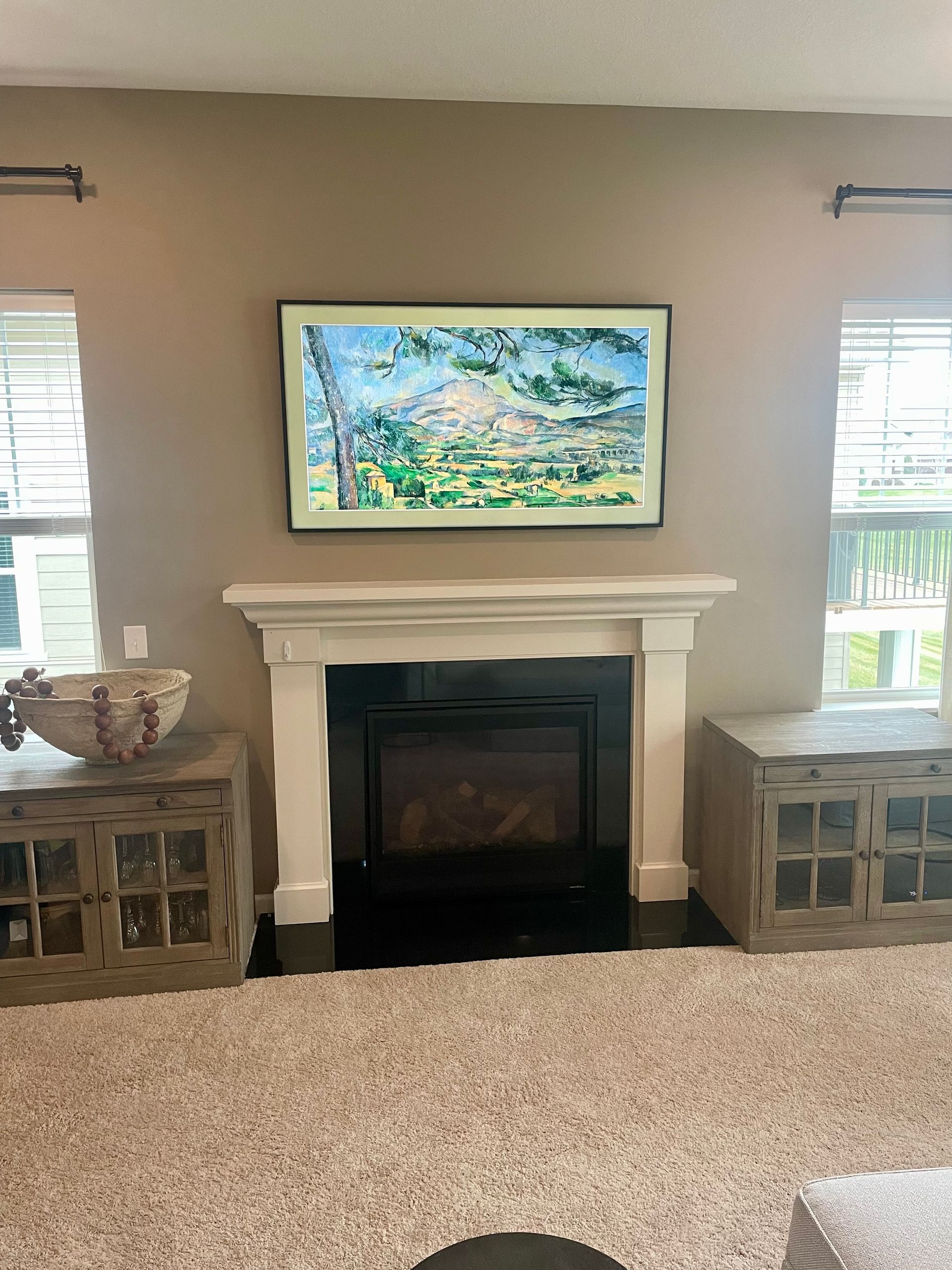 Living room fireplace with white mantel, framed landscape art, and matching side cabinets between two windows
