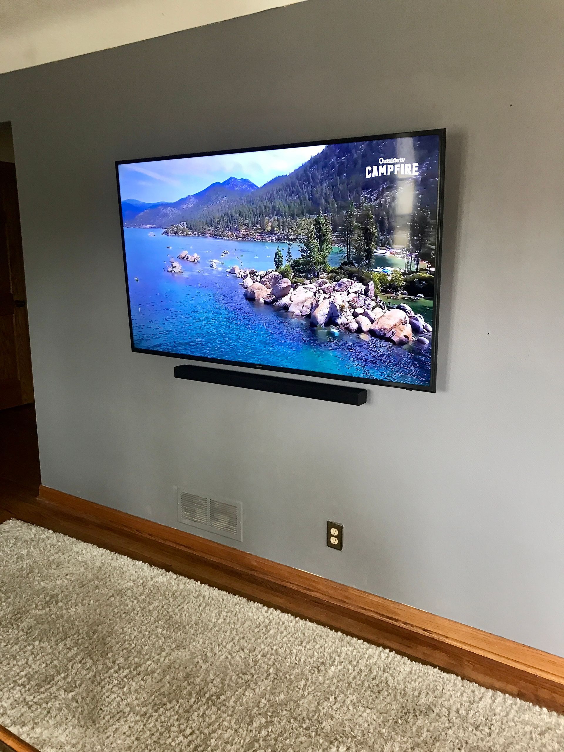 Wall-mounted TV displaying a lake and mountain scene above a soundbar in a carpeted living room.