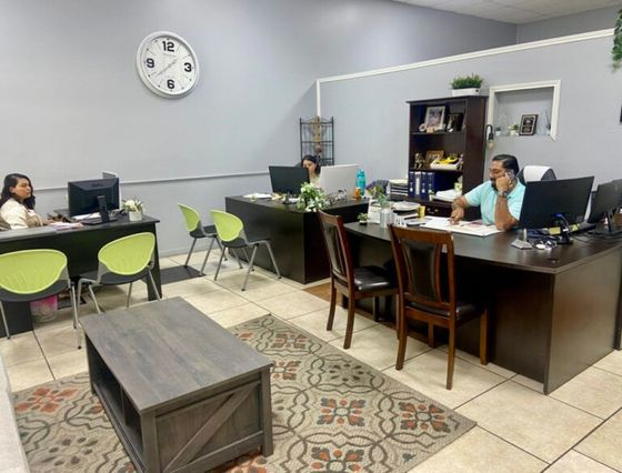 A group of people are sitting at desks in an office with a clock on the wall.