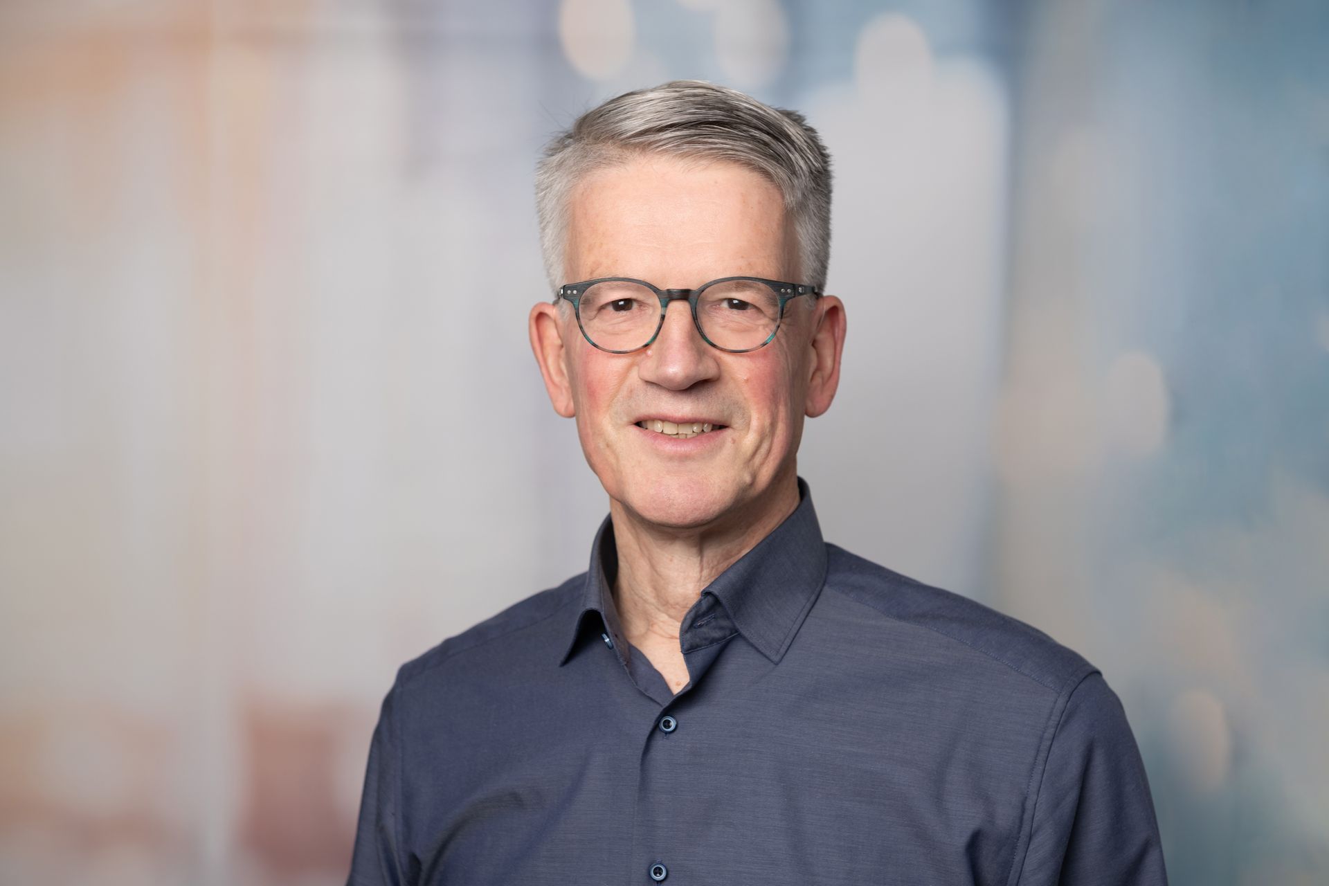 Lächelnder Mann mit Brille und blauem Hemd vor einem leicht verschwommenen Hintergrund