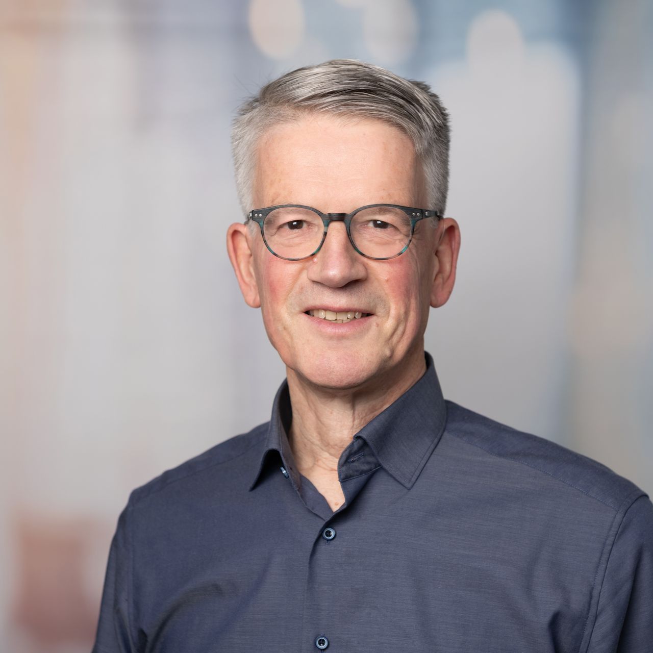 Porträt eines lächelnden Mannes mit Brille und blauem Hemd vor verschwommenem Hintergrund