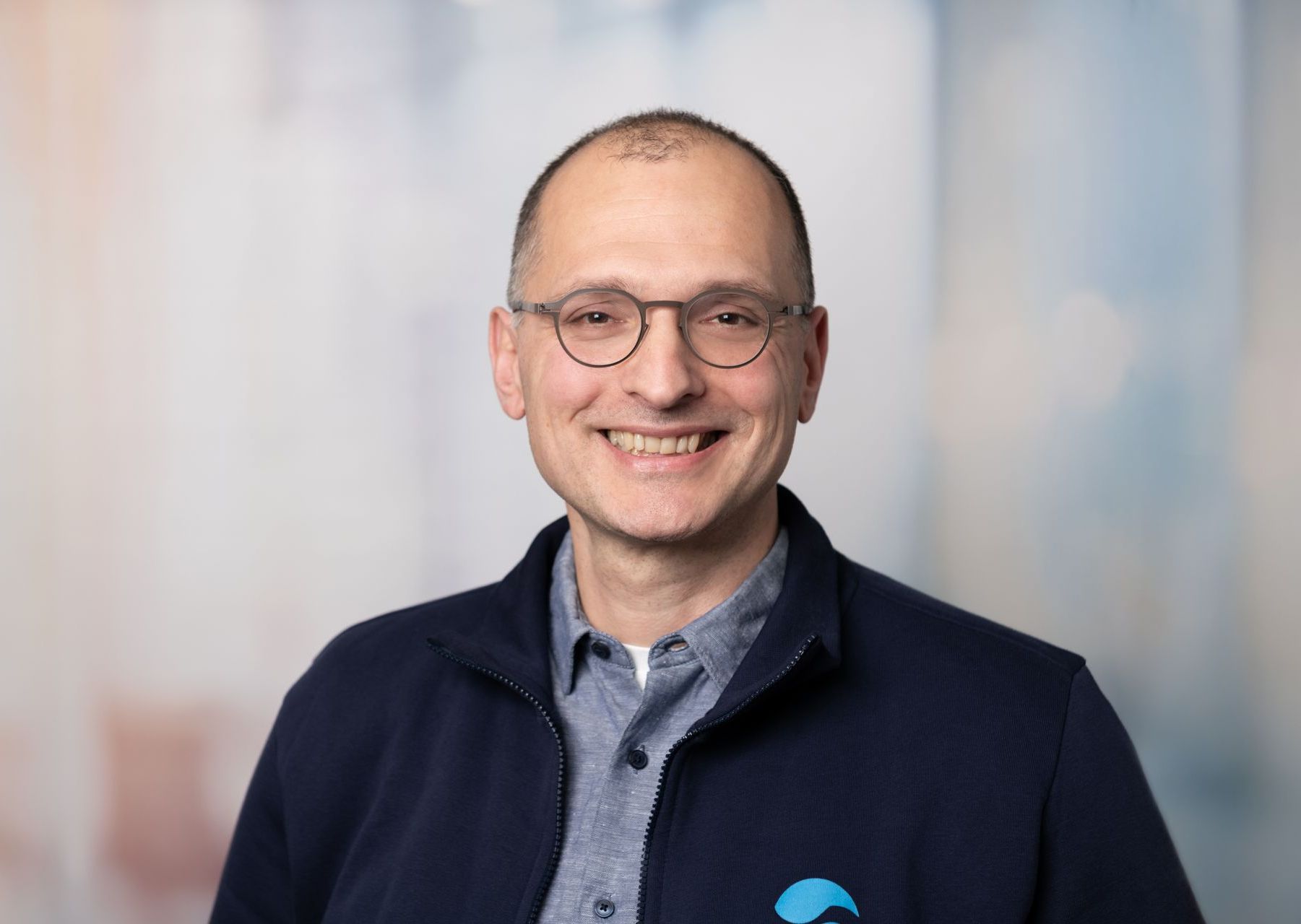 Lächelnder Mann mit Brille und dunkelblauer Jacke vor einem weich verschwommenen Hintergrund
