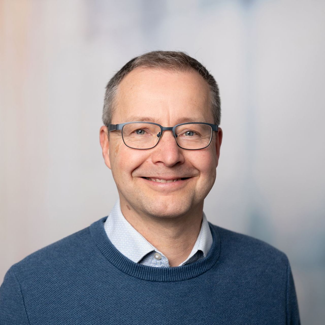Lächelnder Mann mit Brille und blauem Pullover vor hellgrauem Hintergrund