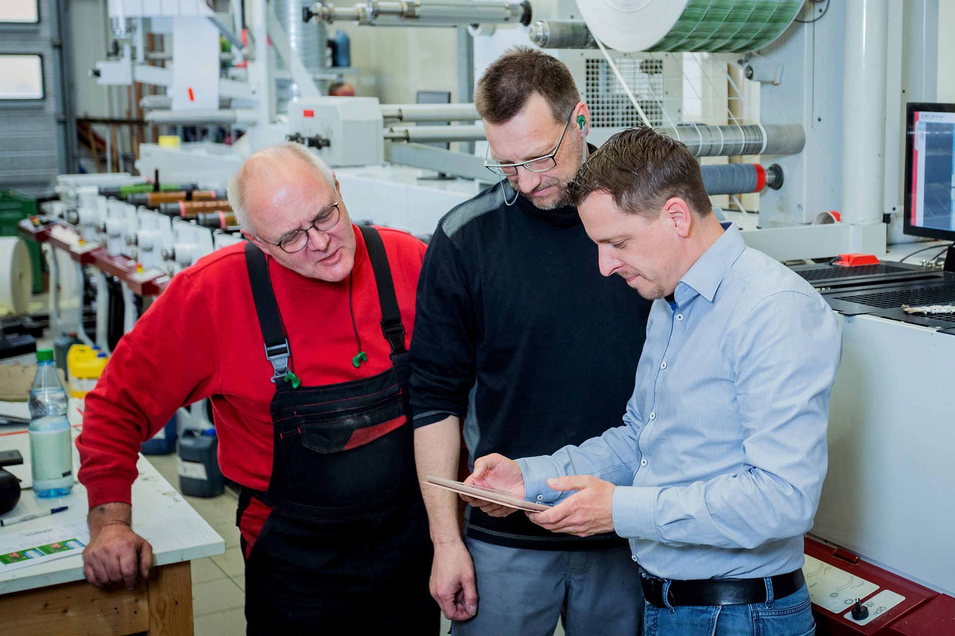 Drei Männer betrachten gemeinsam in einer Werkstatt inmitten von Industrieanlagen ein Tablet.