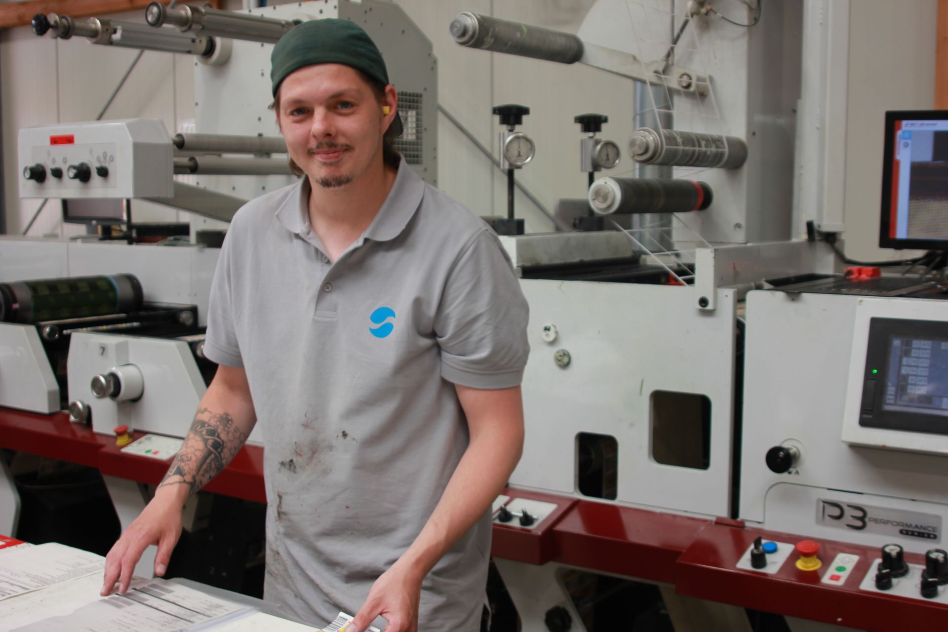 Ein Mann steht in einer Druckerei neben einer großen Druckmaschine. Er trägt ein graues Polohemd und eine grüne Kappe.