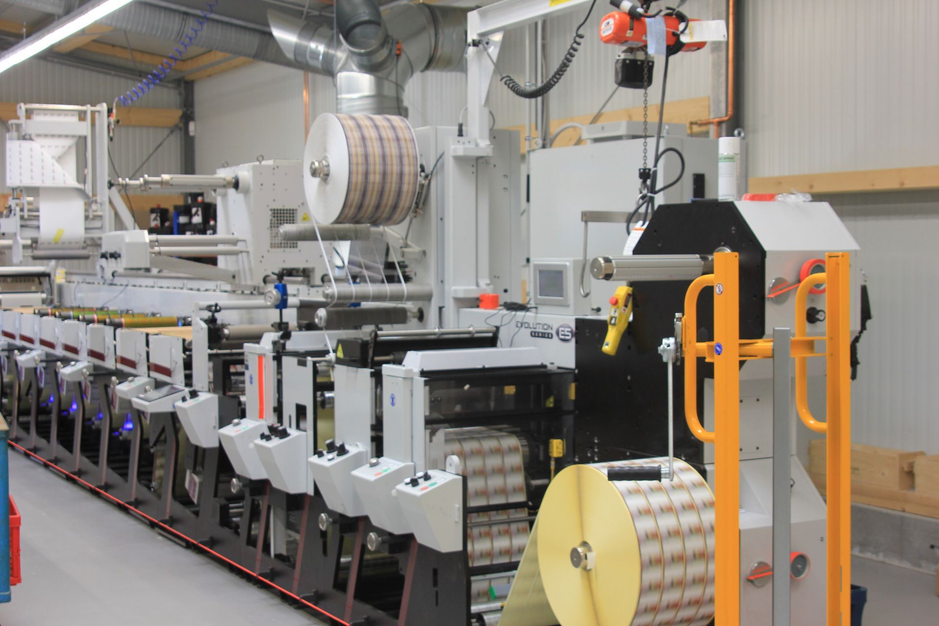 Industrielle Druckmaschine in einer Fabrik, mit großen Papierrollen und orangefarbenen Sicherheitsgeländern.