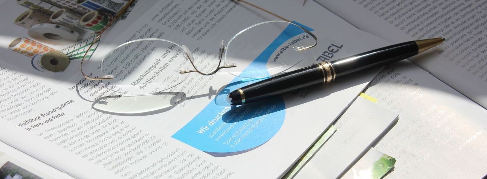 Schwarzer Stift auf einem offenen Notizbuch über einem blauen Haftzettel, daneben Skizzen und Papiere.