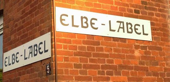 Ecke eines Backsteingebäudes mit zwei weißen Schildern mit der Aufschrift „ELBE-LABEL“
